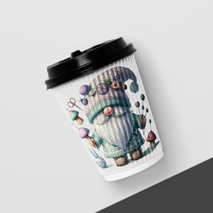 May include: A white paper coffee cup with a black lid features a colorful illustration of a gnome wearing a hat with buttons and holding a mushroom. The gnome is surrounded by other mushrooms, yarn, and a pair of scissors.