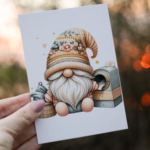 May include: A gnome wearing a knitted hat with floral accents sits next to a small laundry machine. The gnome is holding a basket of clothes.