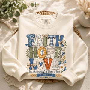 Puede incluir: Una sudadera de cuello redondo color crema con un gráfico de estilo patchwork que muestra las palabras FAITH, HOPE y LOVE. El diseño incluye patrones florales, de cuadros y sólidos en azul, verde y naranja, con lazos, hojas y el texto 1 CORINTHIANS 13:13.