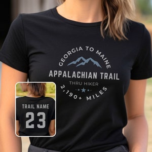 May include: Black t-shirt with a white graphic design that says "Georgia to Maine Appalachian Trail Thru Hiker 2,190+ Miles". The graphic also features a blue mountain range. A separate image shows the back of a black t-shirt with the text "Trail Name 23" in white.