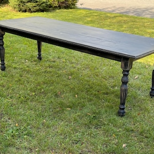May include: A long, rectangular black table with turned legs. The table is made of wood and has a dark gray surface. The table is set on a grassy lawn.