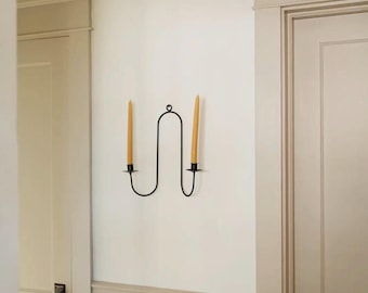 May include: A black metal wall sconce with two yellow candles. The sconce has a unique, curved design and is mounted on a white wall. The candles are slender and taper towards the top, providing a warm, inviting glow.