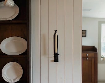 May include: A black metal wall sconce holding a cream-colored candle is mounted on a white paneled wall. A dark wooden cabinet with white plates and a cup is visible on the left.