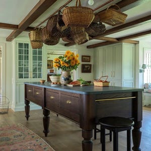 May include: A large dark wood kitchen island with a gray countertop, brass hardware, and turned legs. Above the island, multiple woven wicker baskets hang from a copper rack. A ceramic pitcher filled with yellow sunflowers sits on the surface.