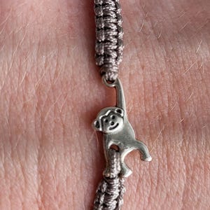 Könnte beinhalten: Ein Makramee-Armband mit einem silbernen Affenanhänger. Das Armband besteht aus grauem und beigefarbenem gewebtem Kordel. Der Affenanhänger hängt am Armband.