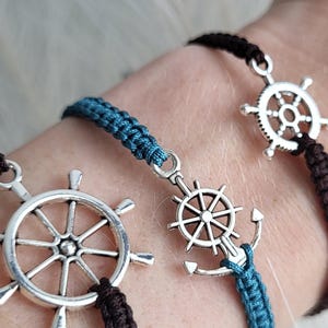 May include: Three nautical-themed bracelets. Each bracelet features a silver-colored ship's wheel charm. One bracelet has a blue woven cord, while the other two have dark brown cords. The bracelets are displayed on a wrist against a white, fluffy background.