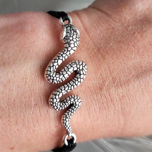 Op de afbeelding: Een zilverkleurige slangenarmband met een gedetailleerd, gestructureerd ontwerp. De slang is het middelpunt, bevestigd aan een zwart koord. De armband wordt om een pols gedragen, met een zachte, vage achtergrond.