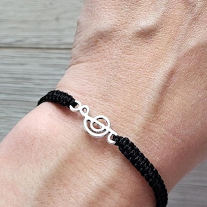 May include: Black woven bracelet featuring a silver-colored treble clef charm. The bracelet is worn on a wrist, with several rings on the fingers and purple nail polish. The background is a light wood grain.