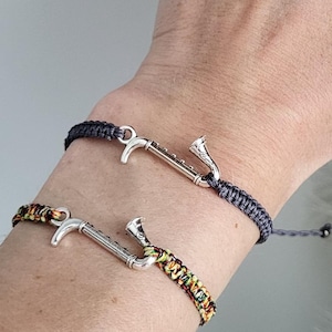 Op de afbeelding: Twee macramé armbanden met zilverkleurige bedels worden getoond op een pols. Eén armband is donkerpaars, de andere is goud en bruin. De achtergrond heeft een zachte, veervormige textuur. De nagels van de persoon zijn geverfd met paarse glitter.