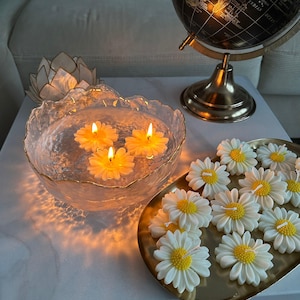 Könnte beinhalten: Dekorative Anordnung mit schwimmenden Blumenkerzen in einer Glasschale. Auf einem goldenen Tablett liegen gänseblümchenförmige Kerzen. Ein Globus und ein lotusförmiger Kerzenhalter tragen zur ruhigen Atmosphäre bei.