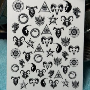 May include: White sticker sheet featuring various occult symbols in black. Designs include goat skulls, all-seeing eyes within triangles, crescent moons, pentagrams, and geometric shapes. The sticker sheet has a slightly rounded edge.