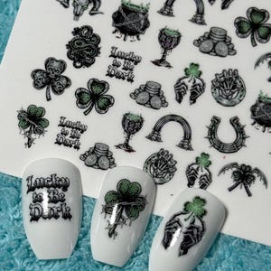 Könnte beinhalten: Nagelkunst-Aufkleber mit verschiedenen gotischen und St. Patrick's Day-Motiven. Enthält Totenköpfe, Kleeblätter, Hände und den Text "Lucky to be Dark". Drei weiße Nägel mit passenden Designs sind ebenfalls sichtbar.