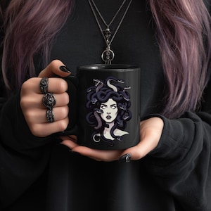 Può includere: Tazza in ceramica nera con un'illustrazione di Medusa. L'opera raffigura Medusa con serpenti al posto dei capelli, occhi bianchi e incarnato pallido. La tazza è tenuta da una persona con smalto nero e diversi anelli.