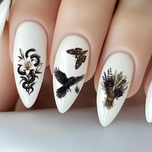 Può includere: Nail art bianco con disegni gotici. Un'unghia presenta un serpente nero attorcigliato attorno a un fiore bianco. Un'altra mostra un corvo nero con un ciondolo e una falena. L'ultima unghia ha un bouquet di rose scure e lavanda.