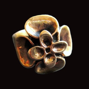 May include: A bronze-colored ring with a floral design. The ring features a central flower with multiple rounded petals and a wide band. The metal has a textured appearance, and the ring is set against a black background.