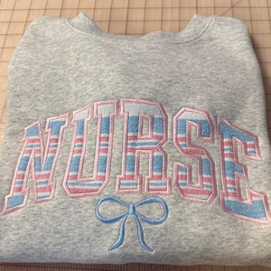 Op de afbeelding: Een grijze sweatshirt met het woord "NURSE" geborduurd in blauwe en roze gestreepte letters. Een blauwe strik is onder het woord geborduurd. De sweatshirt is op een geruit oppervlak gevouwen.