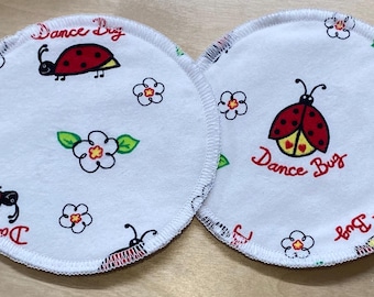 May include: Two white, round nursing pads with a ladybird design. Each pad features a red and black ladybird, the words "Dance Bug" in red script, and floral accents. The pads are made of a soft, absorbent material.