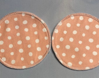 May include: Two round, peach-coloured fabric nursing pads with white polka dots. The pads have a white stitched edge.