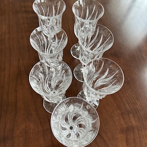 May include: Six clear glass goblets with a decorative, swirling pattern. The goblets are arranged in a circle on a wooden surface.
