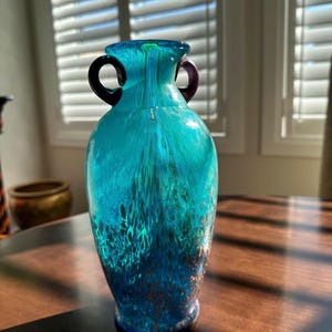 May include: A turquoise glass vase with two dark purple handles. The vase has a textured, layered design with shades of blue and green. The vase sits on a dark wooden surface, with sunlight streaming in from a window in the background.