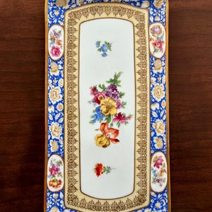 May include: Rectangular porcelain serving tray with a blue floral border and gold trim. The center features a cream-colored panel with a floral bouquet and smaller floral accents. The tray has a gold edge and two small handles.