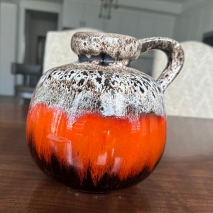 May include: A round, orange, brown and white ceramic vase with a handle. The vase has a mottled glaze with a brown and white pattern on the top half and a solid orange glaze on the bottom half.