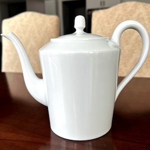 May include: A white porcelain teapot with a curved spout and a large, arched handle. The lid has a small, rounded knob. The teapot is sitting on a dark wooden surface. The teapot is a classic design.