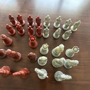 May include: A complete chess set with red and off-white pieces. The red pieces include pawns, rooks, a queen, and other pieces. The off-white pieces include pawns, rooks, a queen, and other pieces. The chess pieces are arranged on a brown surface.