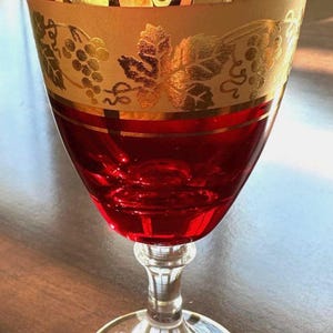 Op de afbeelding: Een robijnrood wijnglas met gouden accenten. Het glas heeft een heldere steel en voet, met een gouden rand. Het bovenste deel van het glas heeft een gouden band met een druivenrankontwerp. Het glas is gevuld met een rode vloeistof.