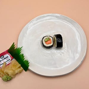 Puede incluir: Dos piezas de sushi en un plato blanco. El sushi está envuelto en alga marina y relleno de arroz, aguacate y salmón. Un pequeño paquete de salsa de soja y una hoja de plástico verde están en la mesa junto al plato.