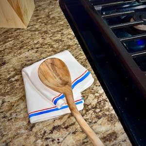 May include: A wooden spoon rests on a folded white kitchen towel with red and blue stripes. The spoon is light brown with a long handle. The towel is on a speckled countertop near a black stovetop.