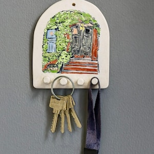 Op de afbeelding: Een keramische sleutelhouder met een gewelfde bovenkant, met een handgeschilderde scène van een deur omgeven door groen. De houder heeft drie pinnen, een sleutelring met sleutels en een lanyard met een clip. De kleuren zijn wit, groen, rood en bruin.