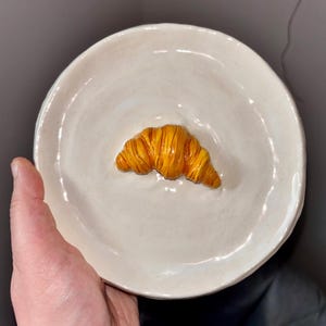 May include: A white ceramic plate with a yellow croissant sculpture on top.