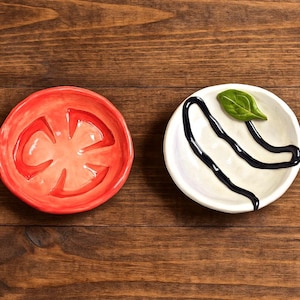 以下が含まれることがあります： 木製の表面に置かれた2つの小さなセラミック皿。1つは赤色で、トマトのスライスのような形をしています。もう1つは白色で、黒いバルサミコグレーズのデザインと緑色のバジルリーフが付いています。前菜の提供に最適です。