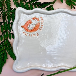 Può includere: Vassoio da portata in ceramica bianca con bordo ondulato. Il vassoio presenta un'illustrazione arancione e bianca di due gatti con le parole "rolling fatties" in corsivo arancione. Foglie di felce verdi sullo sfondo.
