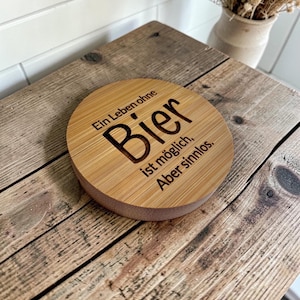 Pode incluir: Um porta-copos redondo de madeira castanho-claro com uma textura de grão natural. A superfície é gravada a laser com o texto em alemão: Ein Leben ohne Bier ist möglich, Aber sinnlos. O porta-copos tem uma superfície superior lisa e plana.
