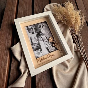 Puede incluir: Un marco de fotos blanco con una imagen en blanco y negro de una pareja. El marco tiene un respaldo de madera y un elemento floral decorativo de madera. El marco de fotos está sobre una superficie de madera con plantas secas y una tela beige.
