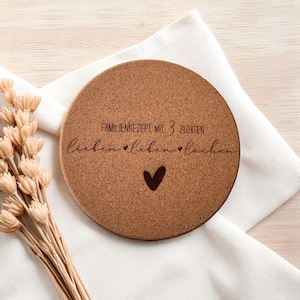 Peut inclure: Un dessous de plat rond en liège avec le texte "FAMILIENREZEPT MIT 3 ZUTATEN lieben lieben lachen" et un symbole de cœur. Le dessous de plat est marron et repose sur un tissu blanc. Des fleurs séchées sont en arrière-plan.
