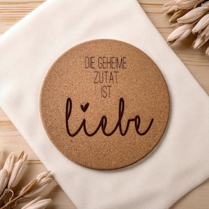 Puede incluir: Un posavasos redondo de corcho con el texto alemán "DIE GEHEIME ZUTAT IST LIEBE" en letras marrones. El posavasos está sobre un paño blanco, con plantas secas en el fondo. El posavasos es de color marrón claro.