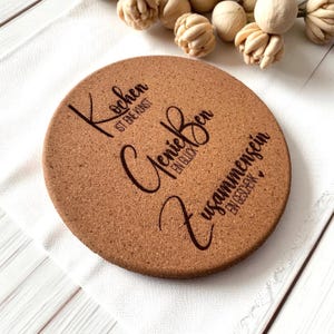 Puede incluir: Un posavasos redondo de corcho marrón con texto marrón oscuro. El texto incluye las palabras "Kochen ist eine Kunst", "GenieBen ein Glück" y "Zusammensein ein Geschenk". El posavasos está sobre un paño blanco.