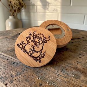 Pode incluir: Um abre-garrafas redondo de bambu com uma ilustração gravada a laser de uma vaca Highland peluda segurando uma garrafa. A peça circular de madeira tem um mecanismo de abertura de metal integrado na parte de trás e um grão de madeira natural.
