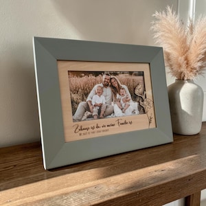 Op de afbeelding: Een grijze rechthoekige fotolijst met een foto van een gezin van vier in een veld. De lichte houten inzet bevat de Duitse tekst "Zuhause ist da, wo meine Familie ist" en "Mit euch ist mein Leben komplett" met een bloemmotief.