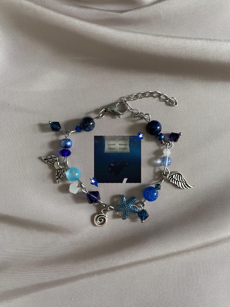 K&ouml;nnte beinhalten: Armband mit blauen Perlen in verschiedenen Formen, darunter runde und facettierte Perlen sowie sternf&ouml;rmige Elemente. Es hat silberfarbene Anh&auml;nger wie Fl&uuml;gel, einen Seestern und eine Spirale. Der Verschluss ist ebenfalls silberfarben.