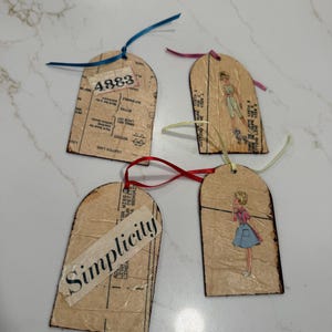 May include: Four arched tags crafted from vintage sewing pattern paper with dark edges. One tag features the number 4883, another displays the word Simplicity, and two tags show illustrations of figures in clothing, each attached with a colored ribbon.