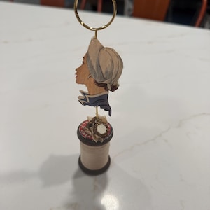 May include: A gold-toned metal wire card holder featuring a circular top loop, mounted on a vintage wooden thread spool. The holder displays a paper cutout of a person in profile wearing a bonnet and a blue collar, with small decorative fabric accents.