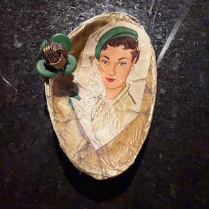 May include: A gold-toned oval dish featuring a decoupage illustration of a person wearing a green beret. Attached to the side of the dish is a cluster of three green buttons and a central metallic gold-toned knot-style button ornament.