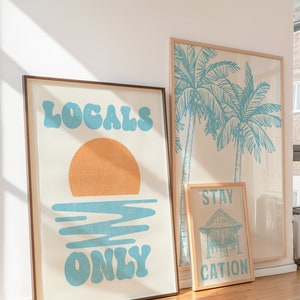 Op de afbeelding: Drie ingelijste prints met een strandthema. De eerste print toont een zonsondergang met de tekst "Locals Only" in blauw. De tweede print toont twee palmbomen in blauw. De derde print toont een klein huis met de tekst "Staycation" in blauw.
