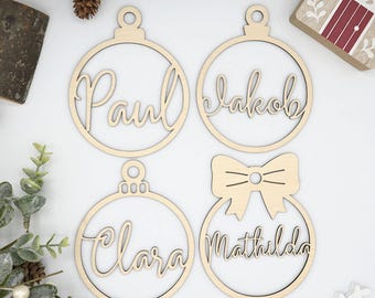 Könnte beinhalten: Vier Weihnachtsbaumkugeln aus Holz mit ausgeschnittenen Namen: Paul, Jakob, Clara und Mathilda. Die Kugeln haben die Form von Christbaumschmuck, eine davon mit Schleife. Sie sind aus hellem Holz gefertigt.