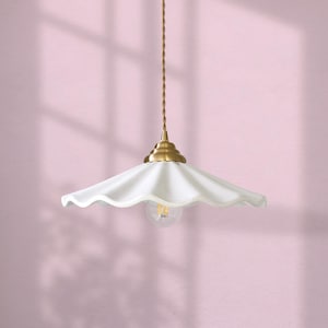 Könnte beinhalten: Eine weiße Pendelleuchte mit gewelltem Rand, Messingbefestigung und sichtbarer Glühbirne. Das Licht hängt an einem gedrehten Goldkabel vor einer rosa Wand. Das Logo "ev lighting" befindet sich unten.