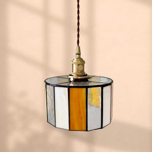 Könnte beinhalten: Hängeleuchte mit einem Lampenschirm aus Buntglas. Der Schirm hat eine geometrische Form mit Glaspaneelen in Weiß, Grau und Gelb. Die Leuchte hat eine messingfarbene Fassung und ein braunes Kabel. Der Hintergrund ist pfirsichfarben.